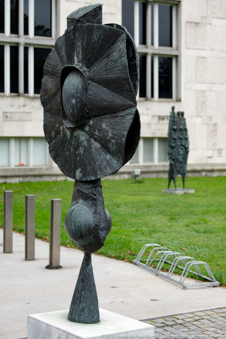 Dark abstract metal sculpture with a broad folded upper form on a tapered pedestal in a museum garden.