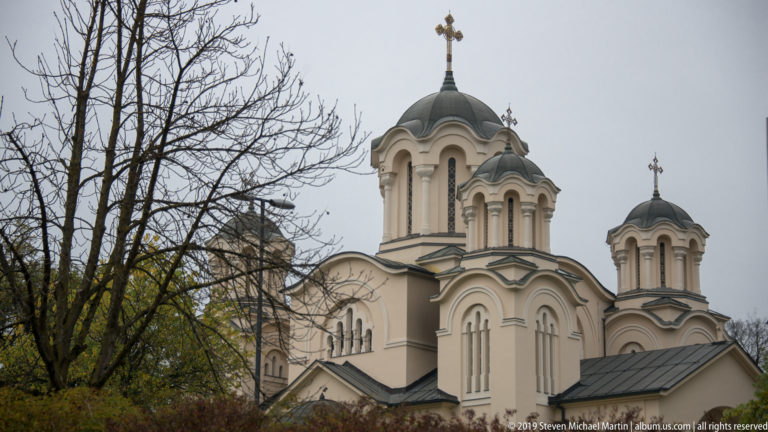 Cream Orthodox church with multiple dark domes and crosses rising behind autumn trees.