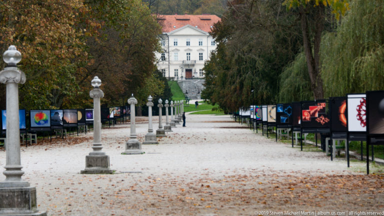 Broad gravel park promenade lined with trees, exhibition panels, lamp posts, and a white building in the distance.