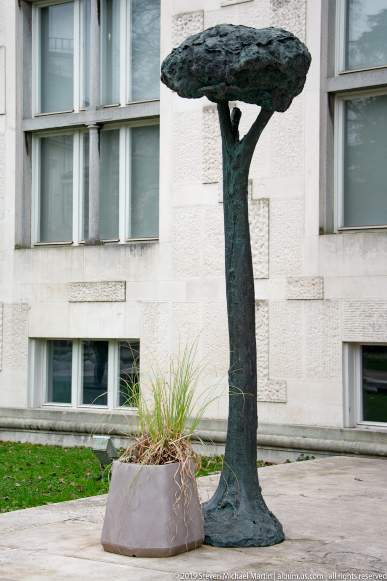 Dark bronze tree-like sculpture with a narrow trunk and broad flattened canopy beside a planter.