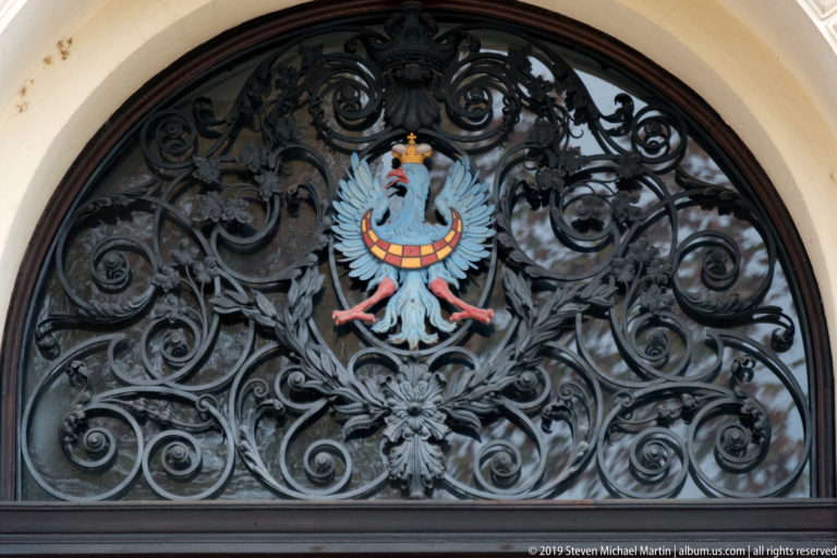 Painted crowned eagle with blue wings and red-and-gold details centered in ornate black wrought-iron scrollwork.