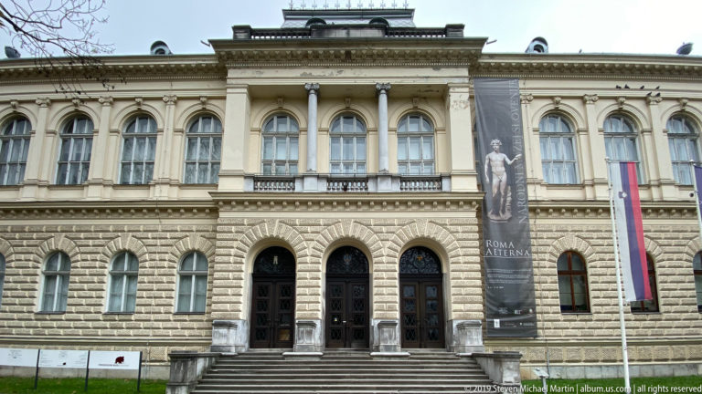 Historic museum façade with rusticated lower story, tall arched windows, Roma Aeterna banner, and Slovenian flag.