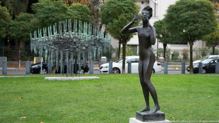 Slender bronze female figure on a plinth with another large abstract sculpture visible behind.