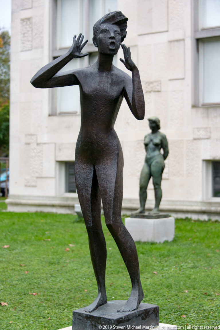Stylized bronze female figure with bent knees and both hands raised beside her face.
