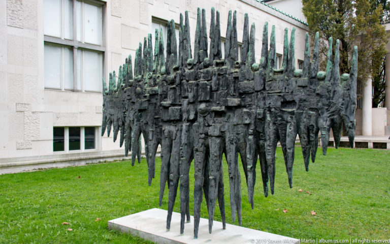 Abstract bronze sculpture composed of many elongated human-like figures clustered into a dense rectangular mass.
