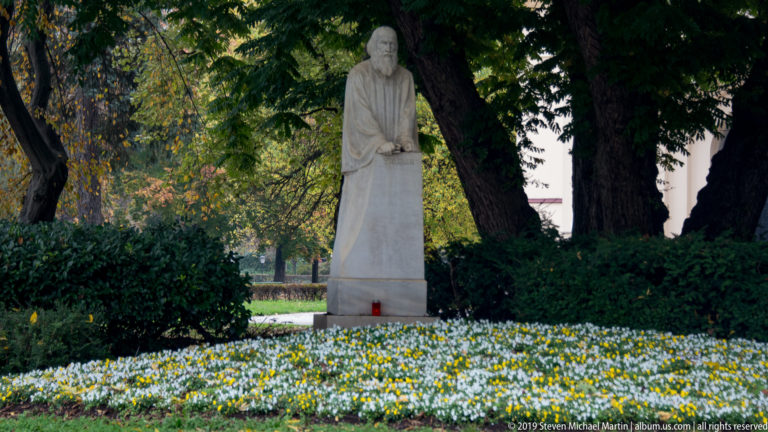 Standing pale stone memorial figure beneath trees, with flowers across the lawn and a red candle at the base.