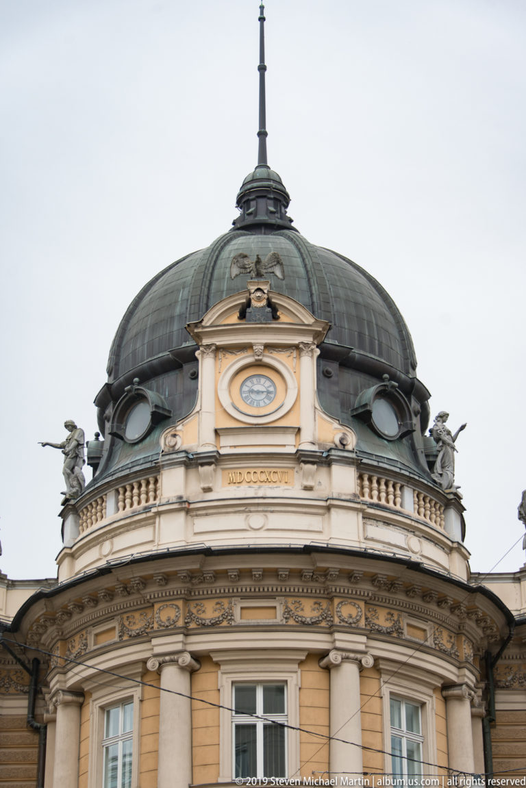 Ornate historic corner building with a dark domed roof, circular upper stage, balustrades, and sculpted figures.