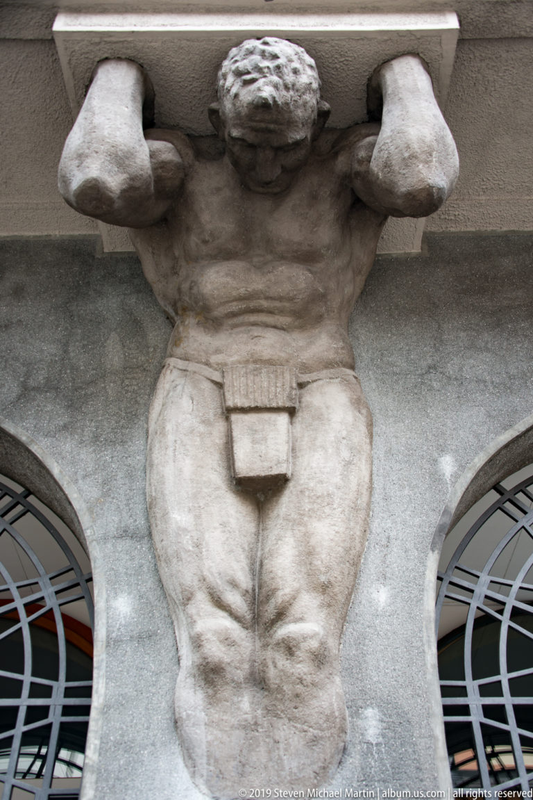 Close-up of a muscular stone atlas figure supporting the Bank of Slovenia façade above an arched window.