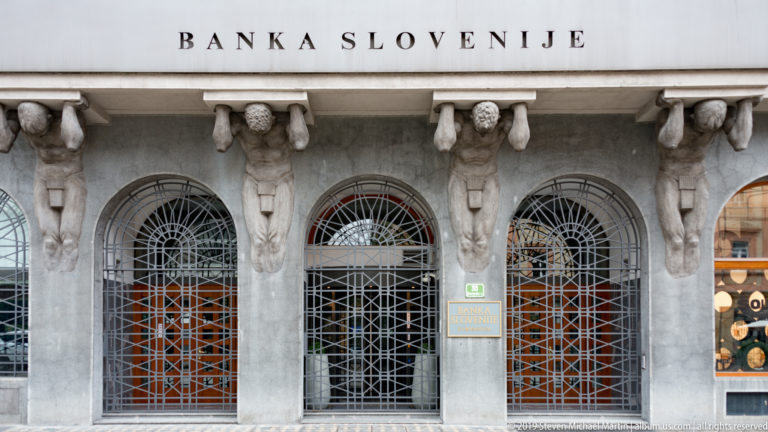 Stone atlas figures supporting the façade above arched, grilled windows of the Bank of Slovenia.