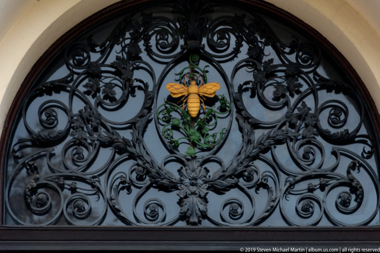 Yellow bee emblem centered in ornate black wrought-iron scrollwork with green painted foliage.