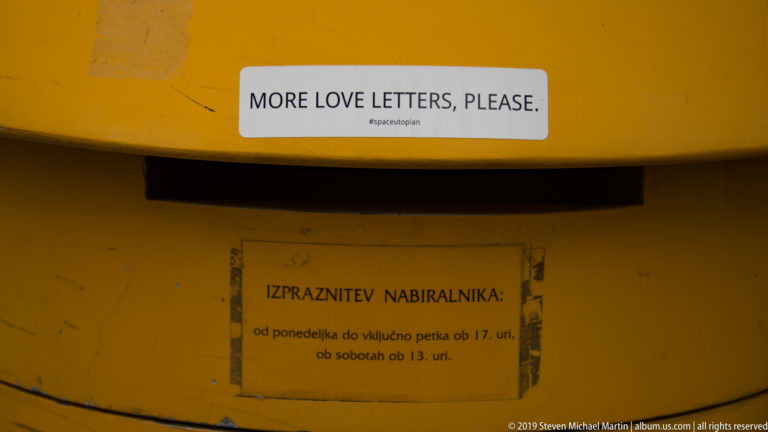Yellow postal box with a sticker reading “MORE LOVE LETTERS, PLEASE.” above a Slovenian collection notice.