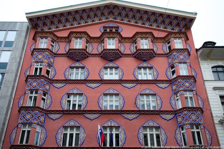 Full pink façade of Vurnik House covered with bright geometric patterns around windows, balconies, and cornices.