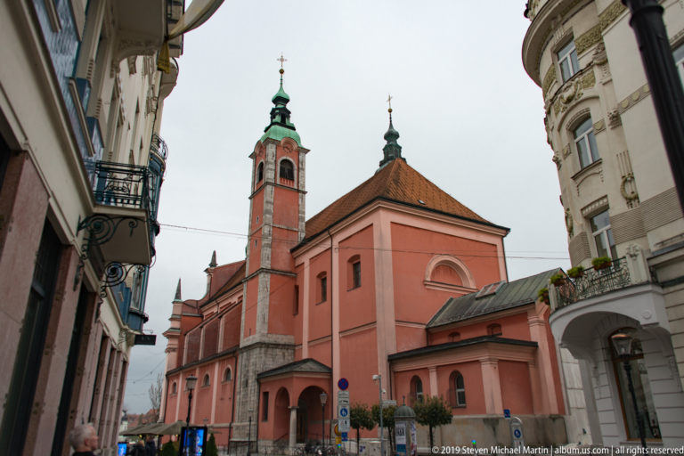 Side view of the red Franciscan Church of the Annunciation with bell tower and adjoining historic buildings.