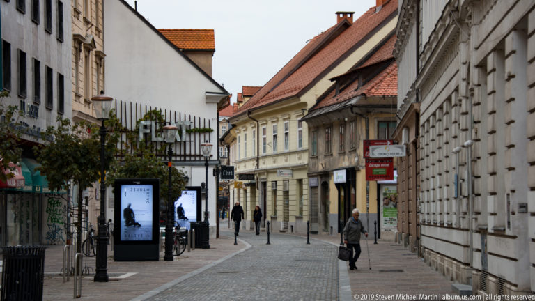 Narrow pedestrian street lined with historic stucco buildings, red-tile roofs, shops, and a few walkers.