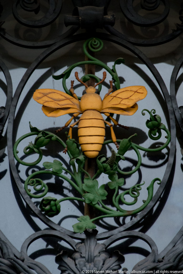 Yellow sculpted bee with wings spread amid green vine motifs in ornate black wrought-iron metalwork.