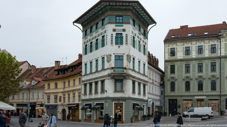 White historic corner building with green shutters, broad eaves, and pedestrians at street level.