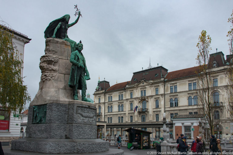 Side view of the Prešeren Monument with historic buildings, pedestrians, and kiosks behind it.
