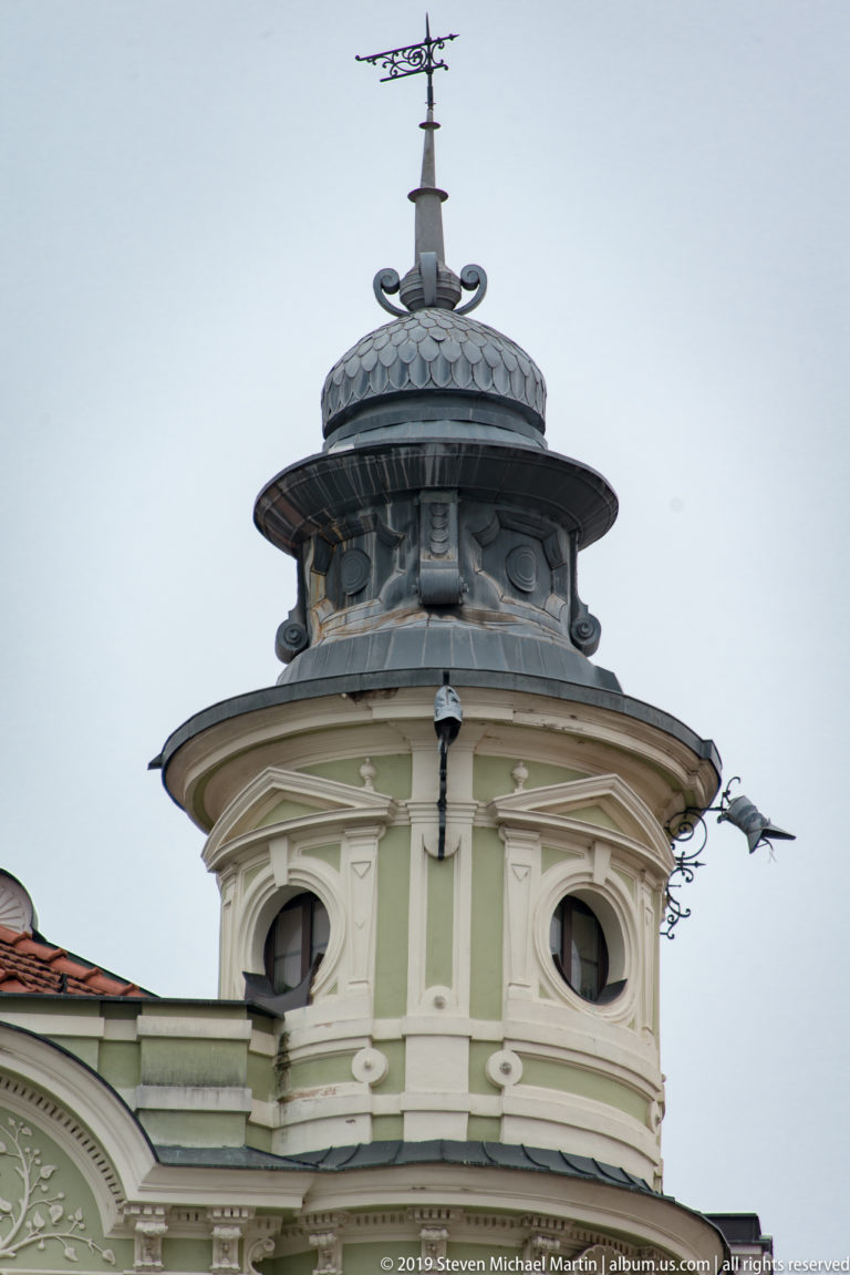 Ornate pale-green corner pavilion with round windows, dark metal roof, and a slender weather vane.