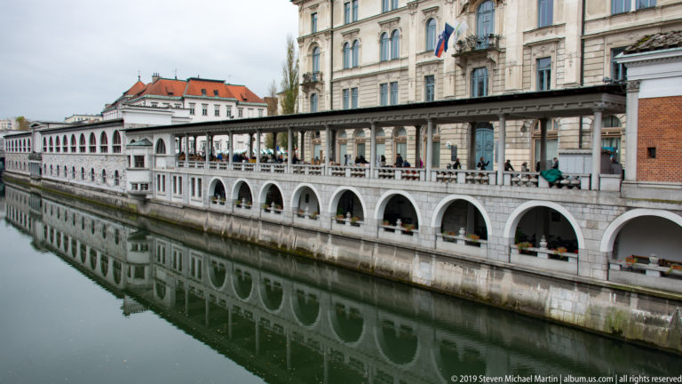 Plečnik’s riverside market colonnade reflected in the Ljubljanica River beneath historic façades.