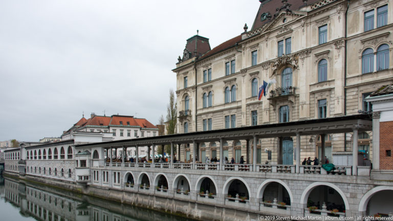 Plečnik market colonnade with repeated arches extending along the Ljubljanica beneath an ornate historic building.