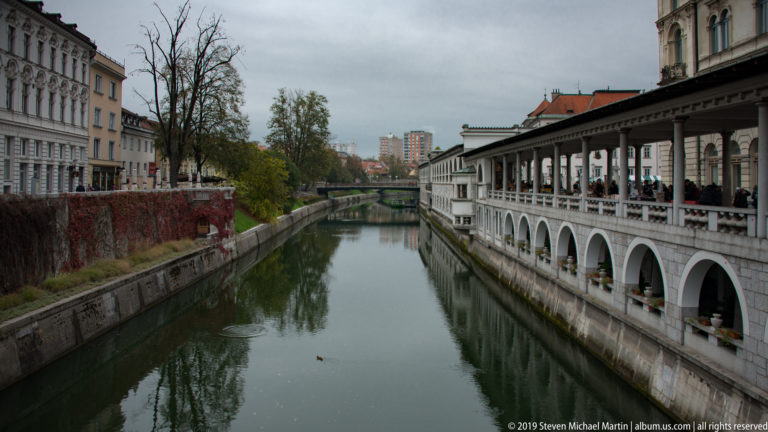 Long arcaded market structure extending beside the Ljubljanica River, with repeated stone arches above the water.