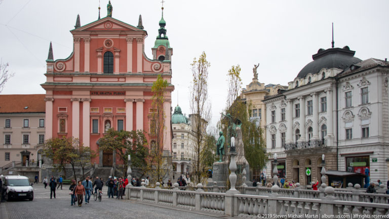 Rose-red Franciscan Church of the Annunciation facing Prešeren Square, with pedestrians and ornate neighboring buildings.