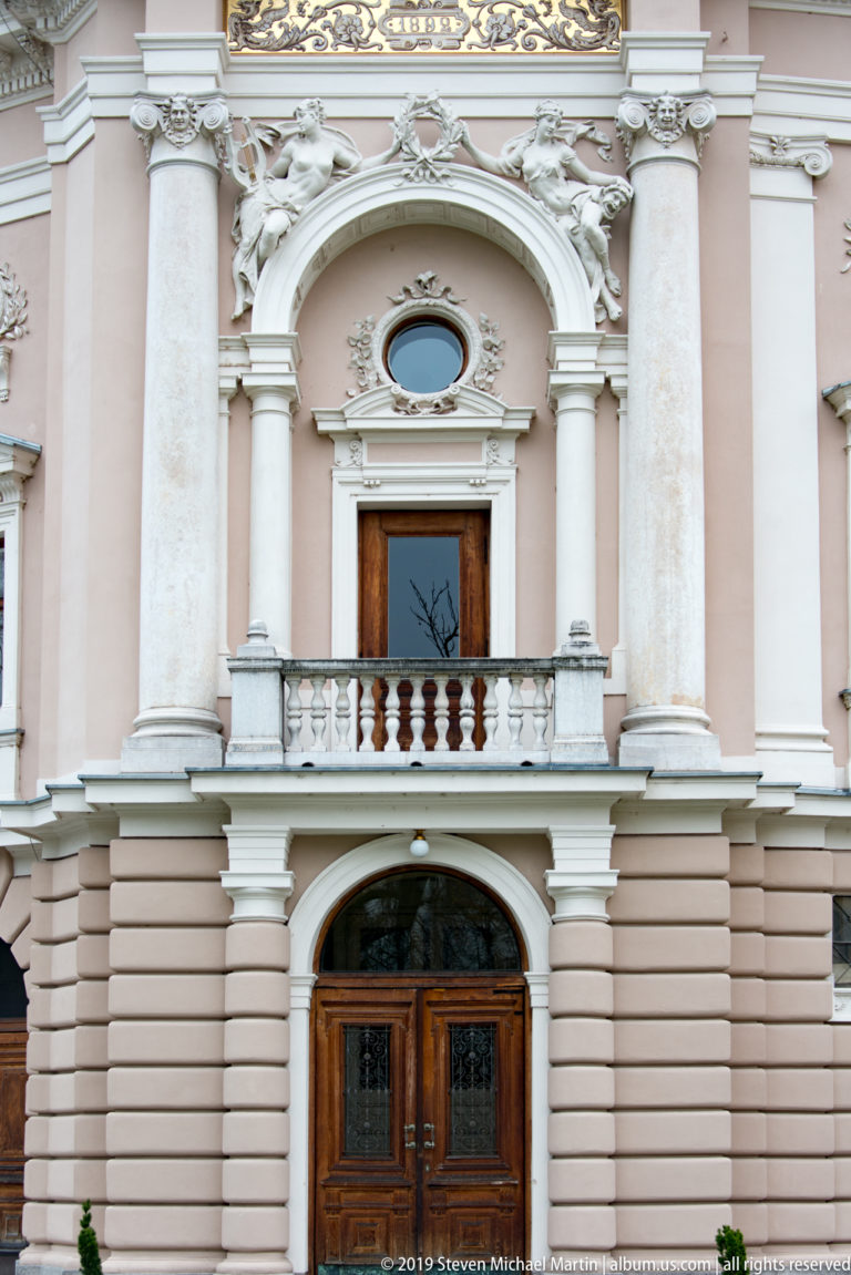Central opera-house bay with wooden entrance, stone balcony, oval window, columns, and sculptural decoration.