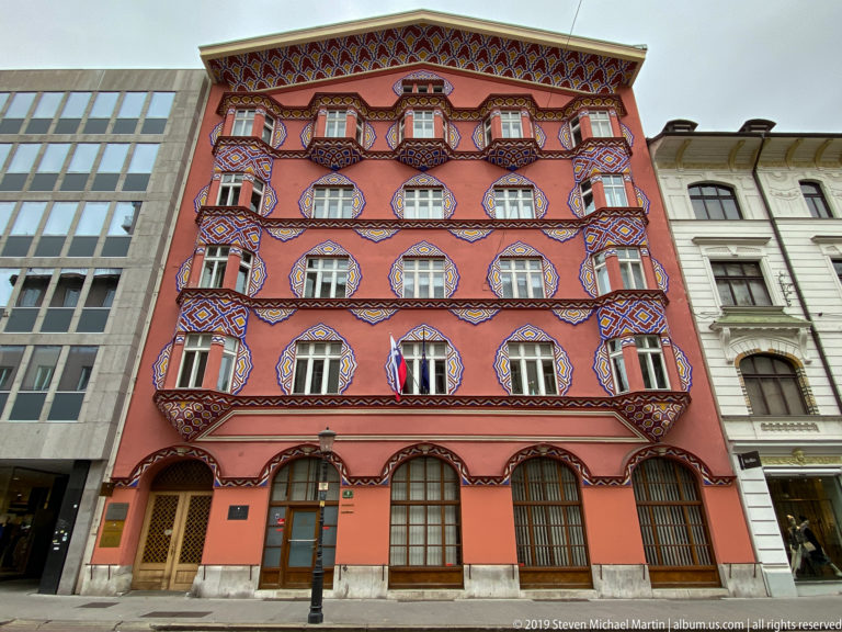 Full red façade of Vurnik House covered with colorful geometric ornament around windows, balconies, and roof eaves.