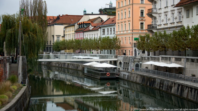 Covered boat on the Ljubljanica River beside stone embankments, trees, cafés, and pastel historic buildings.