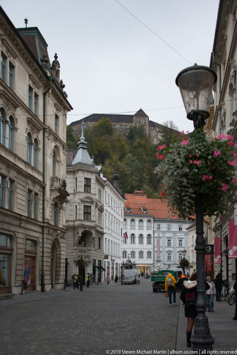 Cobbled Ljubljana street lined with historic buildings, with flower baskets and Ljubljana Castle visible on the wooded hill beyond.