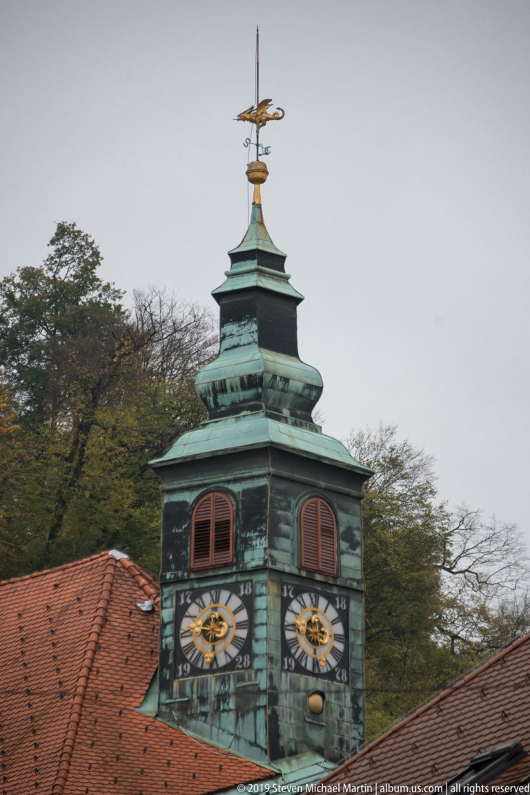 Green copper Town Hall tower with twin clocks and dragon weather vane above red roofs and trees.