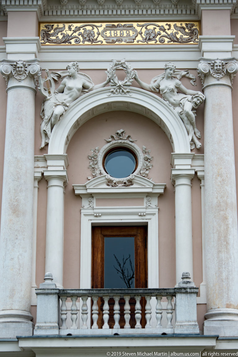 Balustraded balcony and oval window framed by columns and reclining sculptures on Ljubljana’s opera house.