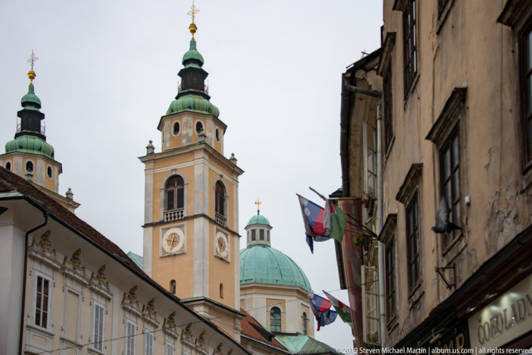 Bell towers and green dome of Ljubljana Cathedral rising above a narrow old-town street with flags and shopfronts.