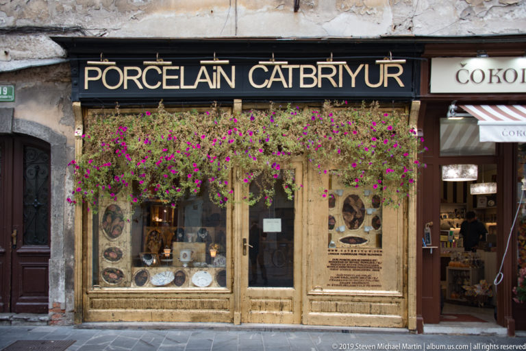 Historic wooden shopfront labeled porcelain, with decorative plates in the windows and flowers hanging above.