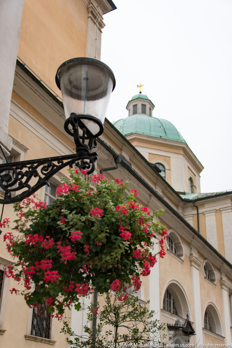 Street lamp and red hanging flowers in front of the green copper dome of Ljubljana Cathedral.