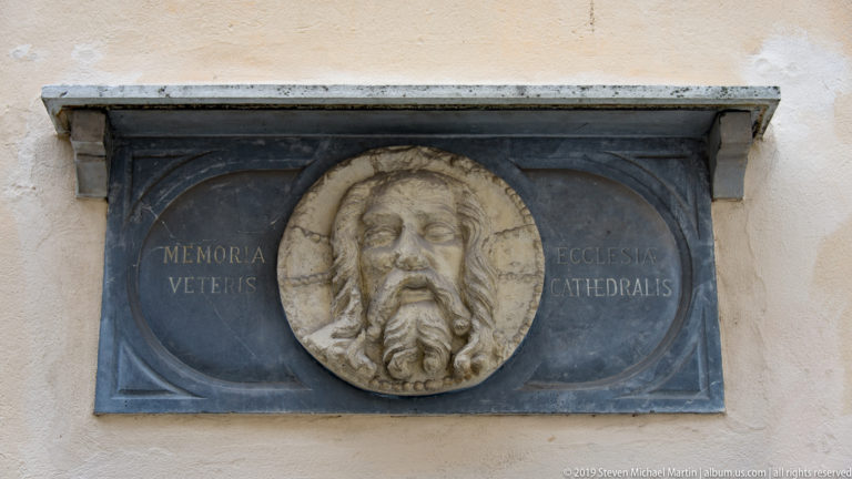 Stone and metal memorial plaque with a sculpted male head and Latin inscription on the cathedral wall.