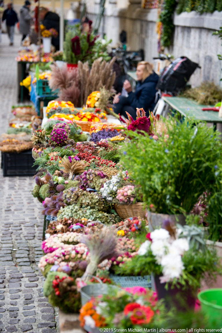 Flower stalls extend across a cobbled market square, with colorful wreaths and blurred figures in the background.
