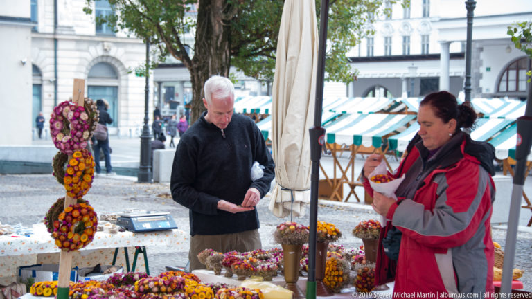Two vendors stand behind an outdoor market stall displaying dried-flower wreaths and handmade decorative goods.