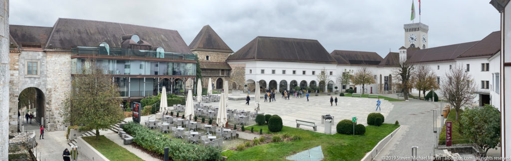 Elevated view across Ljubljana Castle courtyard with historic walls, arcades, café seating, lawns, and visitors.