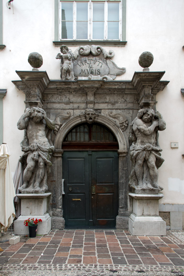 Ornate gray stone doorway with double doors, heraldic carving, and large sculptural figures flanking the entrance.