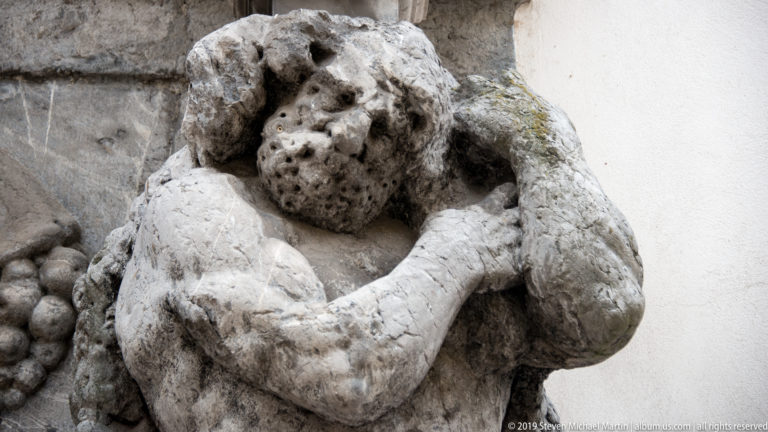 Close view of a weathered stone figure resting its head on one hand, with deeply eroded facial and body surfaces.
