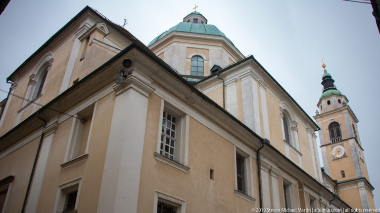 Low-angle view of Ljubljana Cathedral with pale walls, green copper dome, and tall bell tower rising above the street.