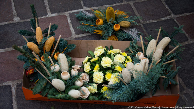 Low floral arrangement with evergreen branches, cream flowers, dried seed pods, and pale elongated decorations in a rectangular container.