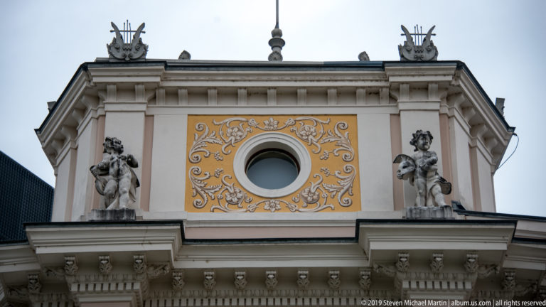 Two stone figures flanking a circular window surrounded by white scrolling ornament on an ocher façade panel.