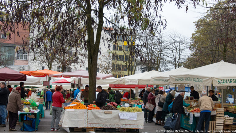 Shoppers and vendors among outdoor produce stalls, umbrellas, and canvas awnings at a Ljubljana market.