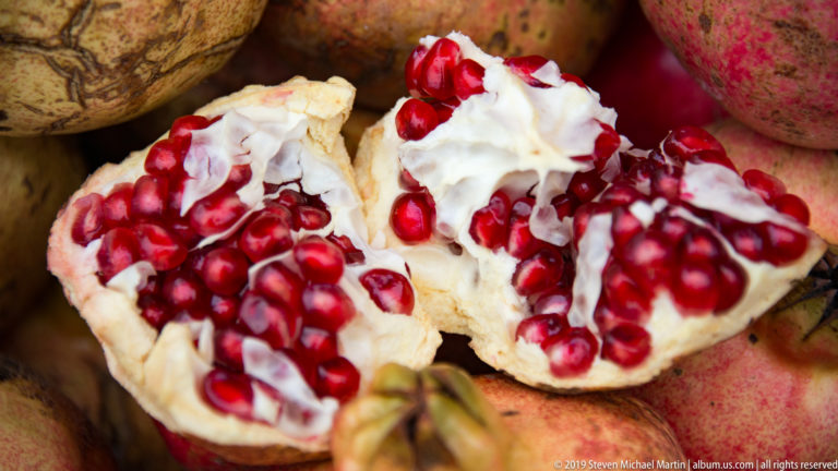 Opened pomegranate showing bright red seeds and white membranes, surrounded by whole fruits at a market stall.