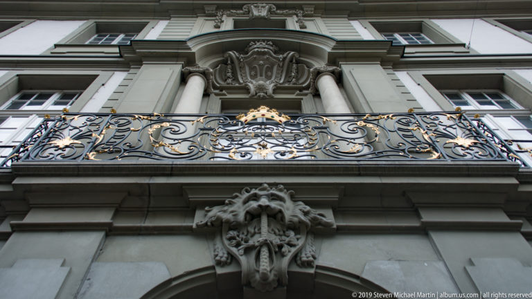 Upward view of a gilded wrought-iron balcony and carved stone façade ornament.