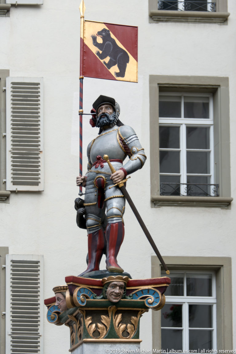 Armored bear fountain figure holding a Bern flag against a pale building façade.