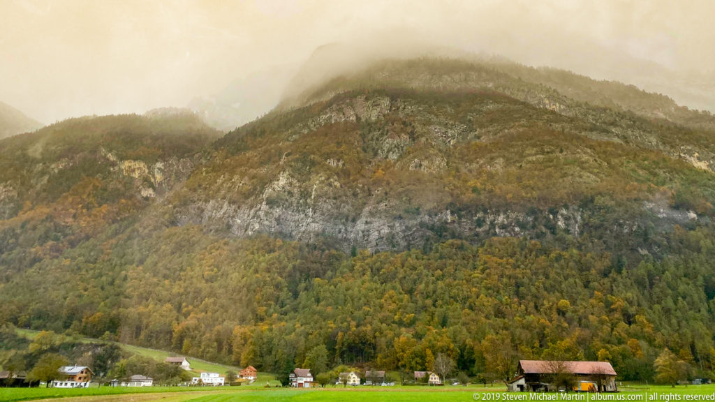 Misty wooded Alpine mountainside with autumn foliage, exposed rock bands, and small houses along the valley floor.