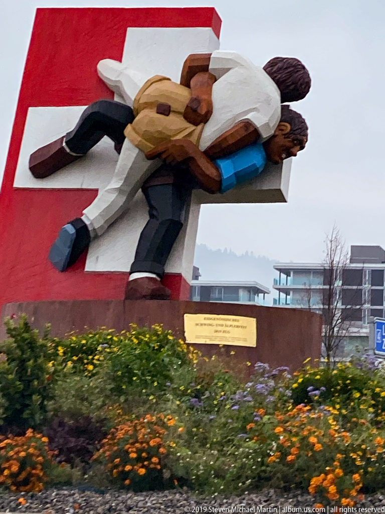 Large stylized sculpture of two embracing human figures mounted on a red roadside structure above flowers.